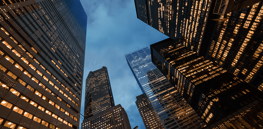 Low-angle upward view of modern city skyscrapers with illuminated office windows against a twilight evening sky.